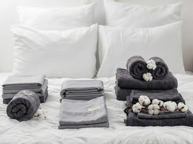 Crisp-white-bedding-and-folded-linens Neatly folded white and grey cotton sheets and towels arranged on a white bed with pillows in the background.